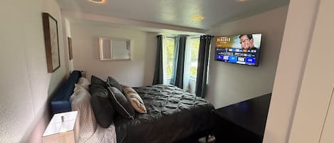Ein Schlafzimmer mit einem Bett, einem an der Wand montierten Fernseher, einem Nachttisch und einem Fenster mit Vorhängen.