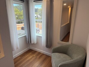 A room with a wooden floor, a grey armchair, and a window with white curtains. - Whisper Valley Villa. New Remodel. Hot Tub! Fenced back yard, High speed WiFi. (Eureka)