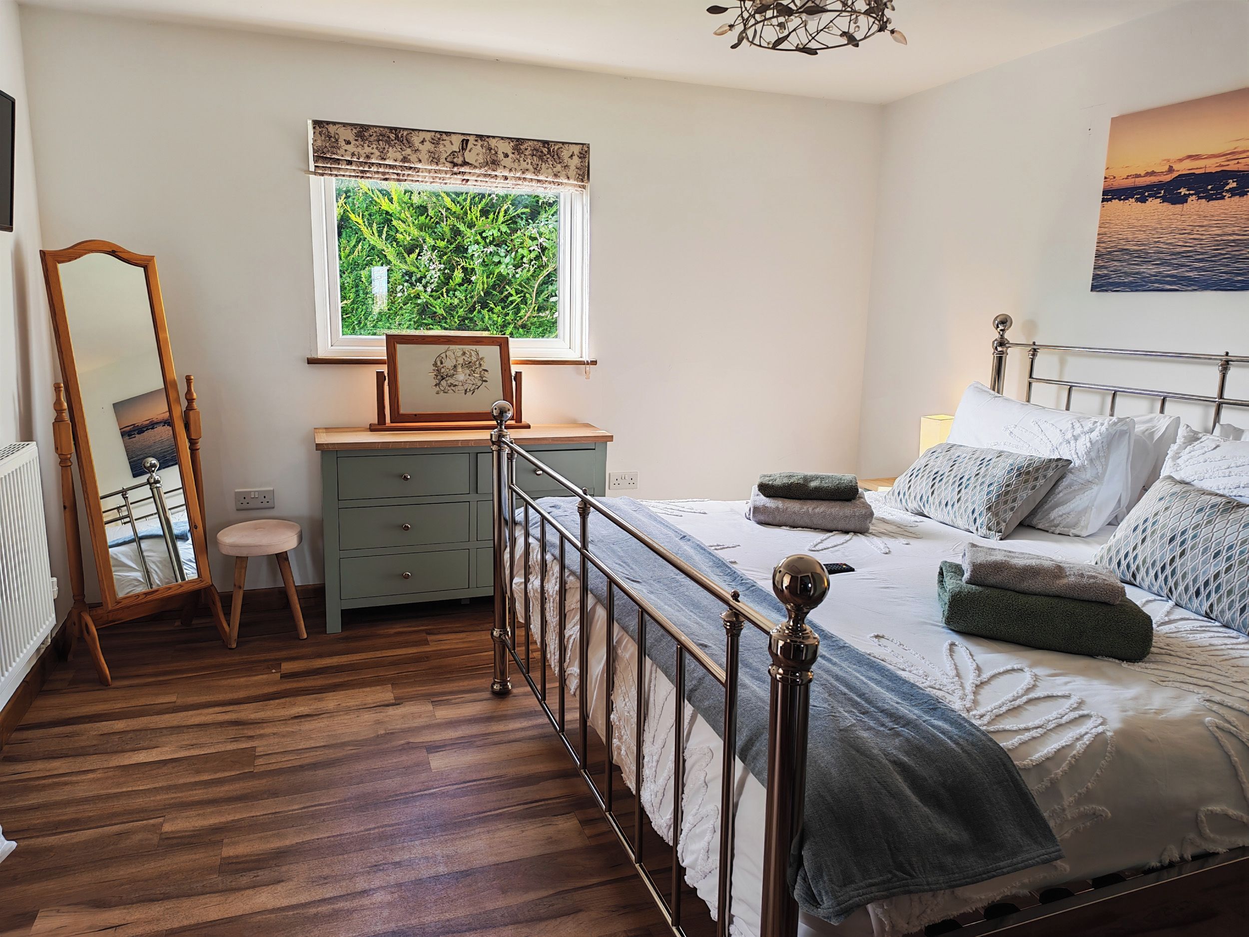 Un dormitorio con piso de madera, una cama con cabecera metálica, un tocador, un espejo y una ventana con vista a la vegetación.