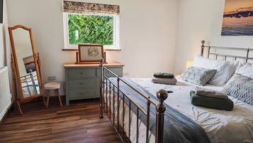 Un dormitorio con piso de madera, una cama con cabecera metálica, un tocador, un espejo y una ventana con vista a la vegetación.