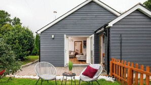 A grey wooden building with a covered patio area featuring two wicker chairs and a small table with a plant.