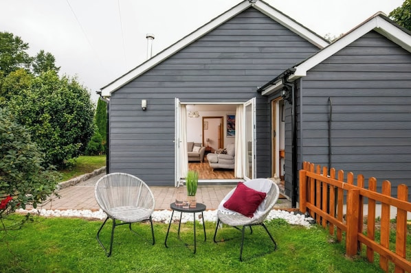 A grey wooden building with a covered patio area featuring two wicker chairs and a small table with a plant. - Willow Lodge (Falmouth)