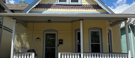 A two-story house with a front porch, a small garden with purple flowers, and a clear blue sky.