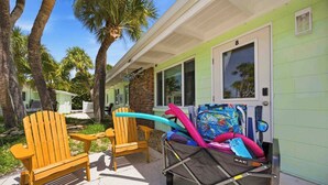A beach house with a patio, two Adirondack chairs, a colorful beach chair, and a small cart with a blue hose. - Pirate's Paradise | Manasota Key Beach Condo | Dock & Pool (Englewood)