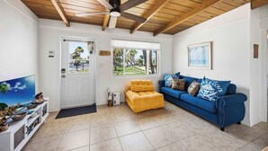 A room with a ceiling fan, a yellow armchair, a blue sofa, and a television on a white stand. - Pirate's Paradise | Manasota Key Beach Condo | Dock & Pool (Englewood)