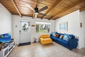 Ein Zimmer mit Deckenventilator, einem gelben Sessel, einem blauen Sofa und einem Fernseher auf einem weißen Podest.