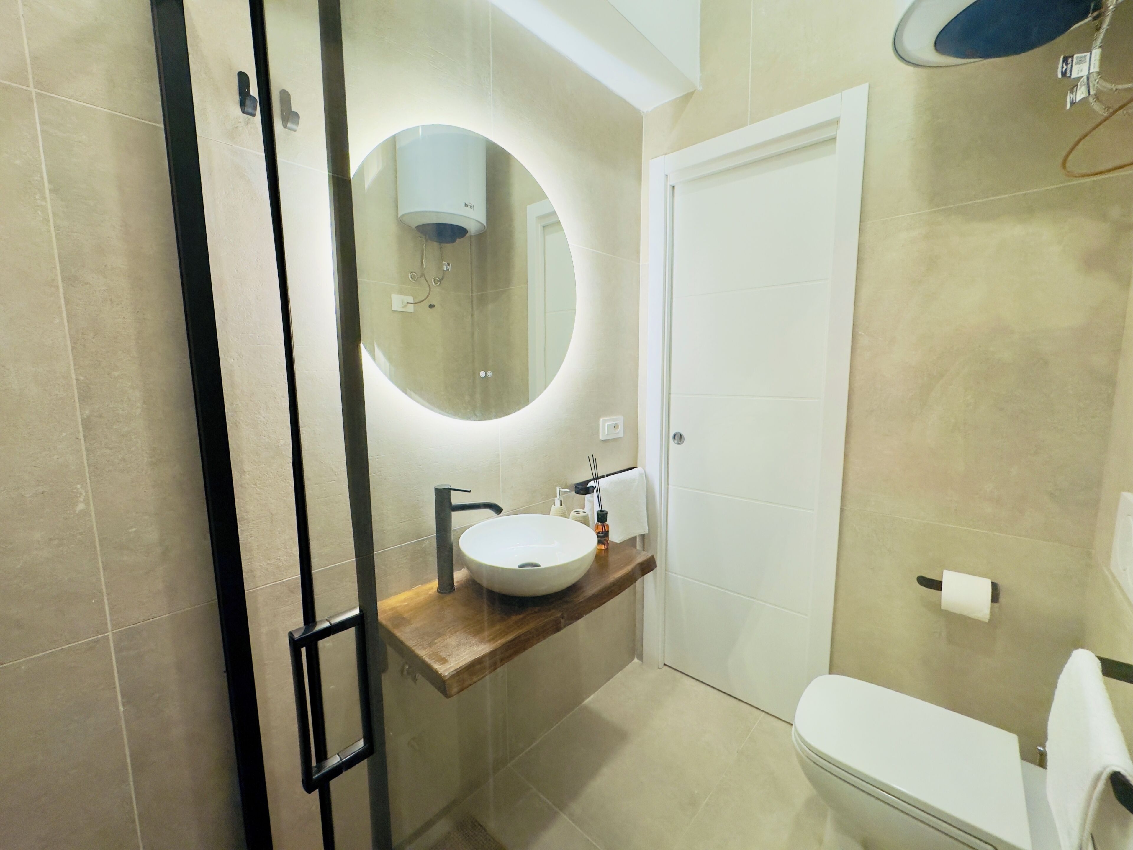 Een moderne badkamer met een douchecabine van glas, een ronde spiegel, een witte wastafel en een toilet.