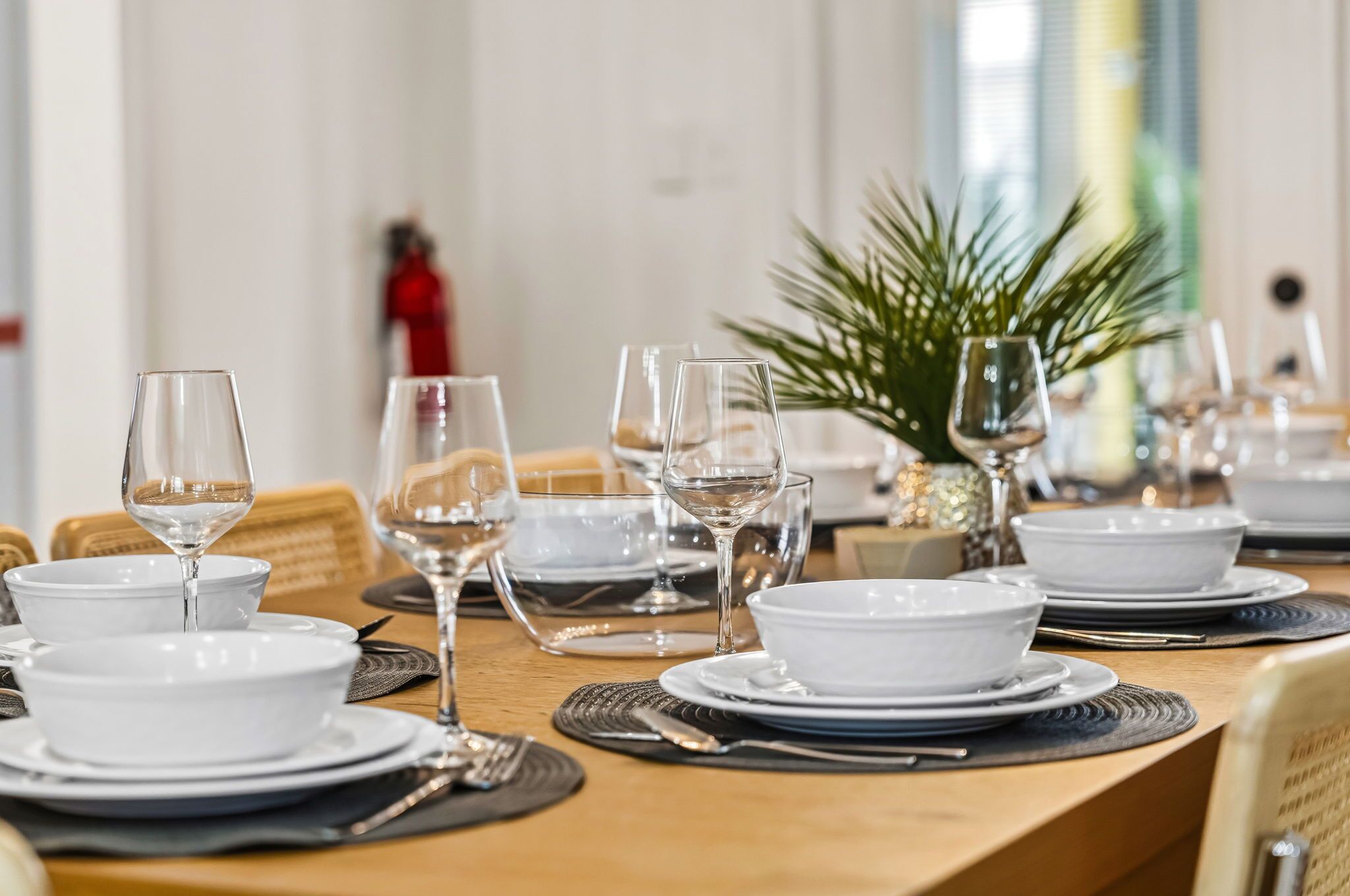 Una mesa de comedor dispuesta con platos, cuencos y copas de vino, con una planta al fondo.