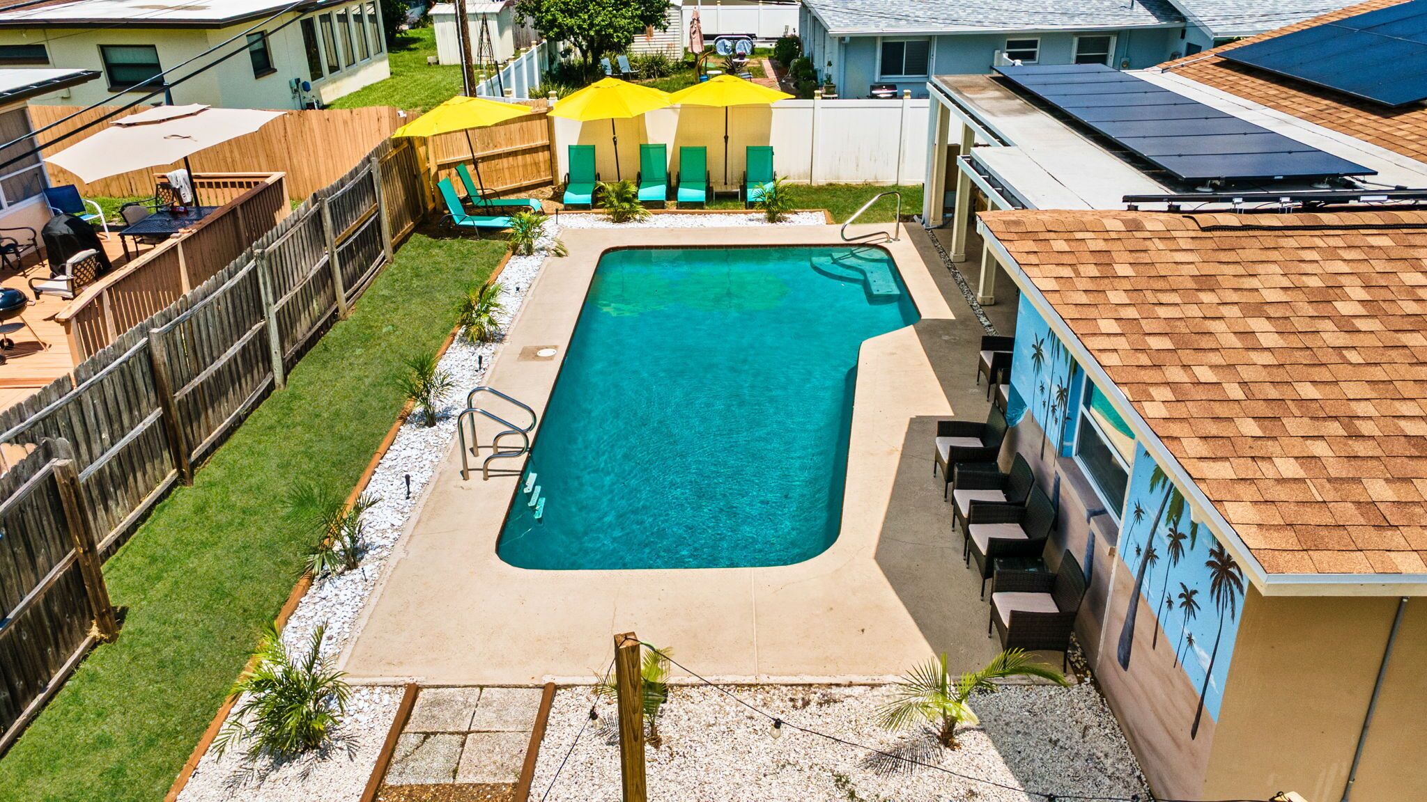 Piscina rectangular con un borde de hormigón, rodeada por una valla de madera y un pequeño jardín con palmeras.