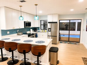 A modern kitchen with white cabinets, a stainless steel refrigerator, and a bar area with stools. - The Tiki Hideaway - Indoor Pool. Tiki Bar. Modern Beach Vibes (Homosassa)
