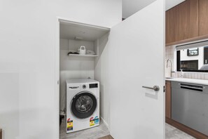 A compact laundry room with a Bosch washing machine, a small shelf with items, and a modern kitchen area with a dishwasher. - Secure Quiet CBD Christchurch Townhouse | Free Parking + EV (Christchurch)