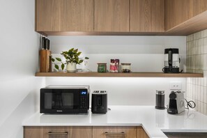 A kitchen with wooden cabinets, a microwave, a toaster, a coffee maker, and a plant on the countertop. - Secure Quiet CBD Christchurch Townhouse | Free Parking + EV (Christchurch)