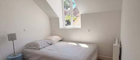 A bedroom with a sloped ceiling, a bed, a bedside table, a lamp, a toilet, and a window.