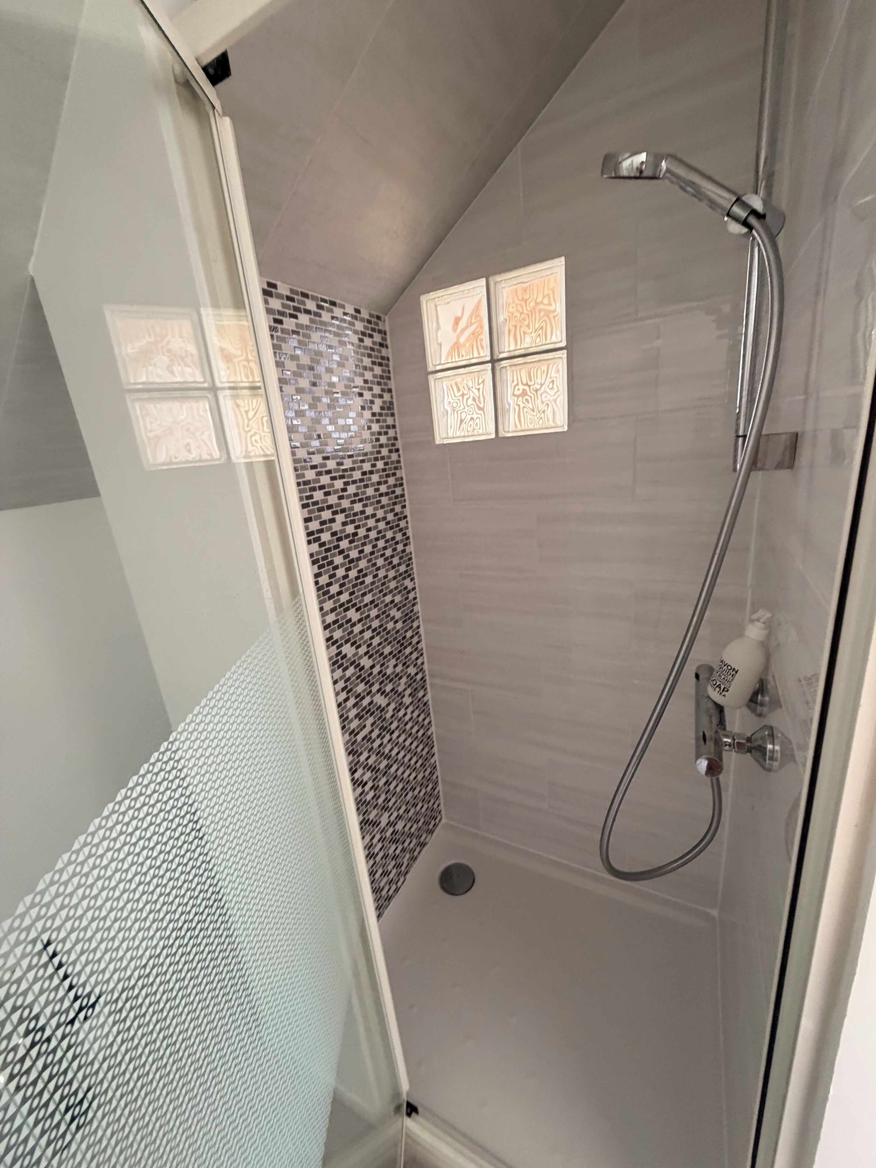 Une douche avec une porte en verre, un pommeau de douche à main et un distributeur de savon fixé au mur.