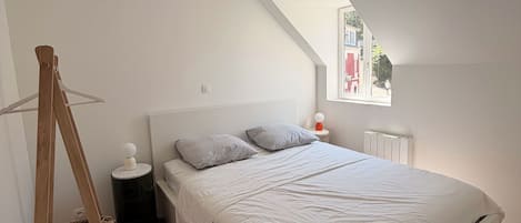 A bedroom with a sloped ceiling, a bed with white bedding, a bedside table with a lamp, and a window with a view of the outside.