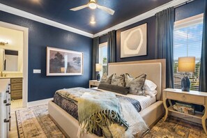 Een slaapkamer met een bed, nachtkastje, lamp, schilderij en een raam met gordijnen.