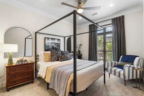 A four-poster bed with a ceiling fan, a bedside table with a lamp, a striped armchair, and a large window with curtains. - The Bentonville Brownstone | Luxury Stay Downtown (Bentonville)