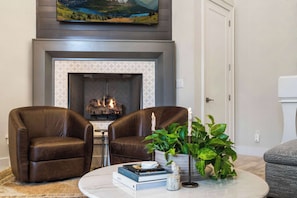 A living room with a fireplace, two leather chairs, a coffee table with books and a plant, and a painting above the fireplace. - The Bentonville Brownstone | Luxury Stay Downtown (Bentonville)
