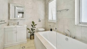 Een badkamer met een bad, toilet, wastafel met kastjes, een spiegel en een potplant.
