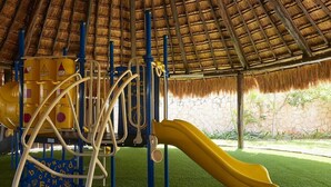 Un'area giochi con tetto di paglia, uno scivolo giallo e strutture per arrampicata.