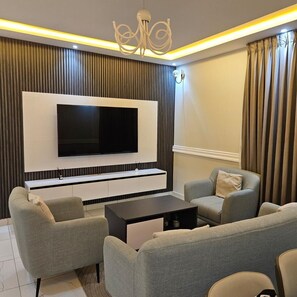 A modern living room with a flat-screen TV mounted on a wall, a black coffee table, and two grey sofas with cushions. - Cozy 1-Bedroom Apartment in Dansoman Control, Enjoy comfort, style and beauty. (Accra)