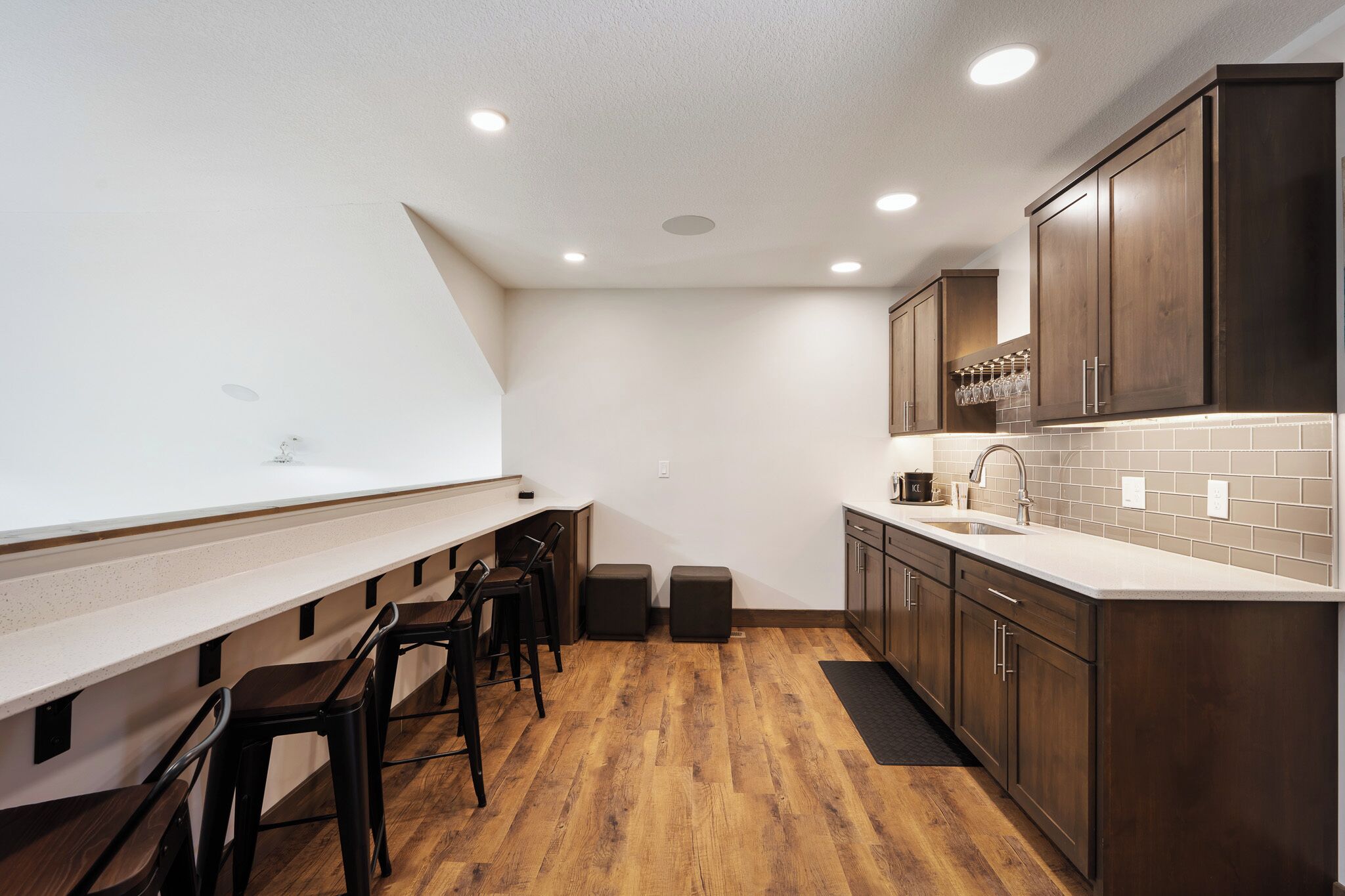 A compact kitchen with a long counter, stools, wooden cabinets, and a tiled backsplash.