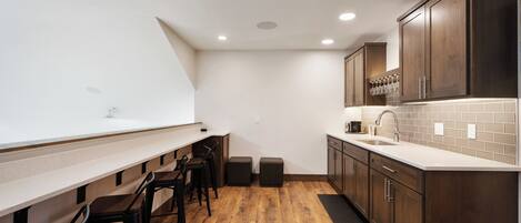 A compact kitchen with a long counter, stools, wooden cabinets, and a tiled backsplash.