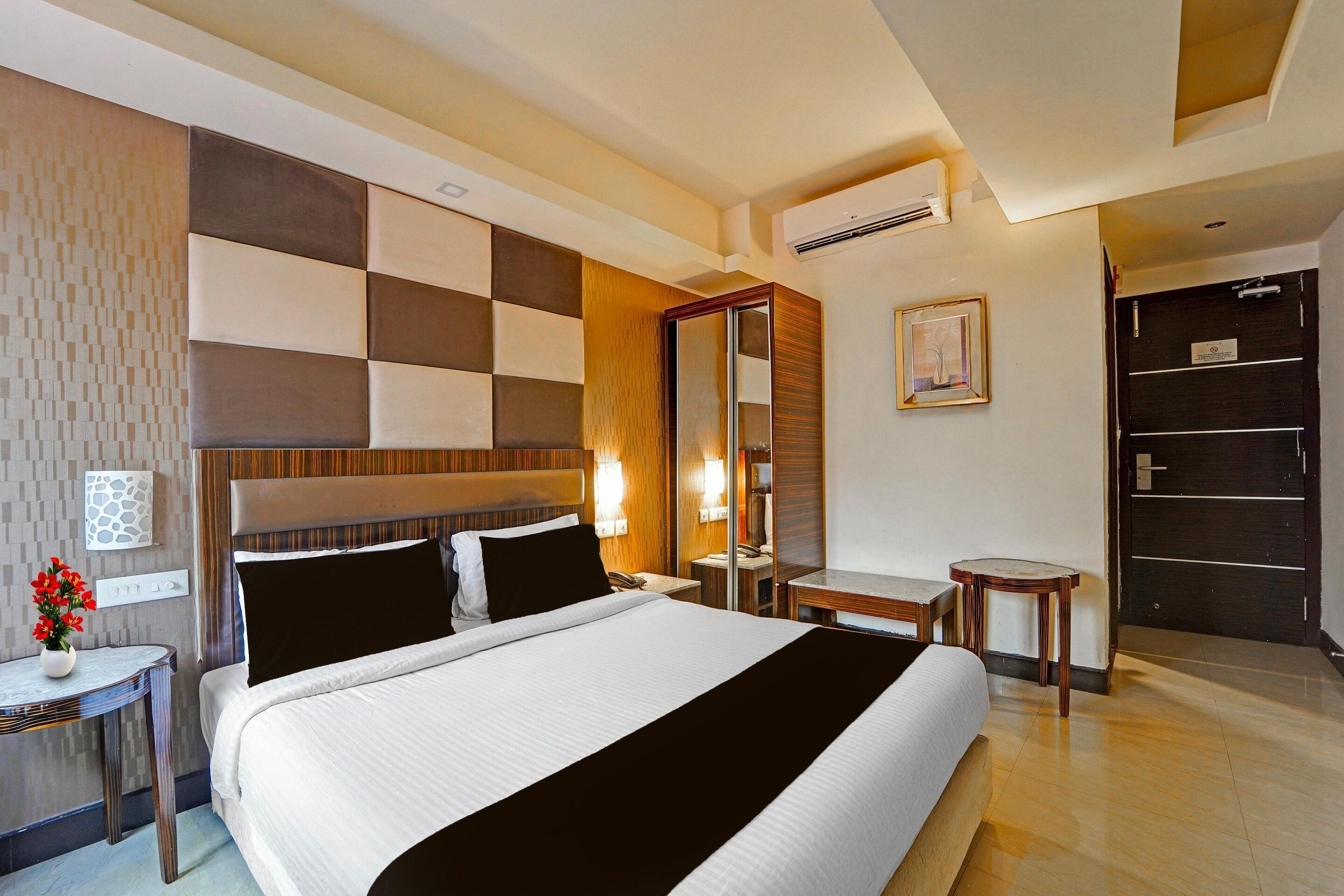 A hotel room with a large bed, bedside tables, a desk, a chair, a mirror, a framed picture, and an air conditioning unit.