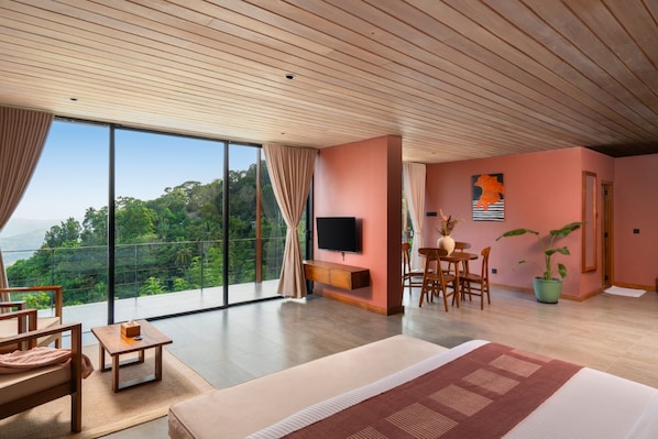 A modern hotel room with a large bed, a TV, a dining area, and a balcony with a scenic view.