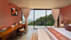 Une chambre avec un grand lit, un bureau avec des livres, une machine à café et une vue sur un paysage montagneux par une porte vitrée.