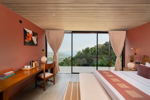 Ein Schlafzimmer mit einem großen Bett, einem Schreibtisch mit Büchern, einer Kaffeemaschine und Blick auf eine gebirgige Landschaft durch eine Glastür.