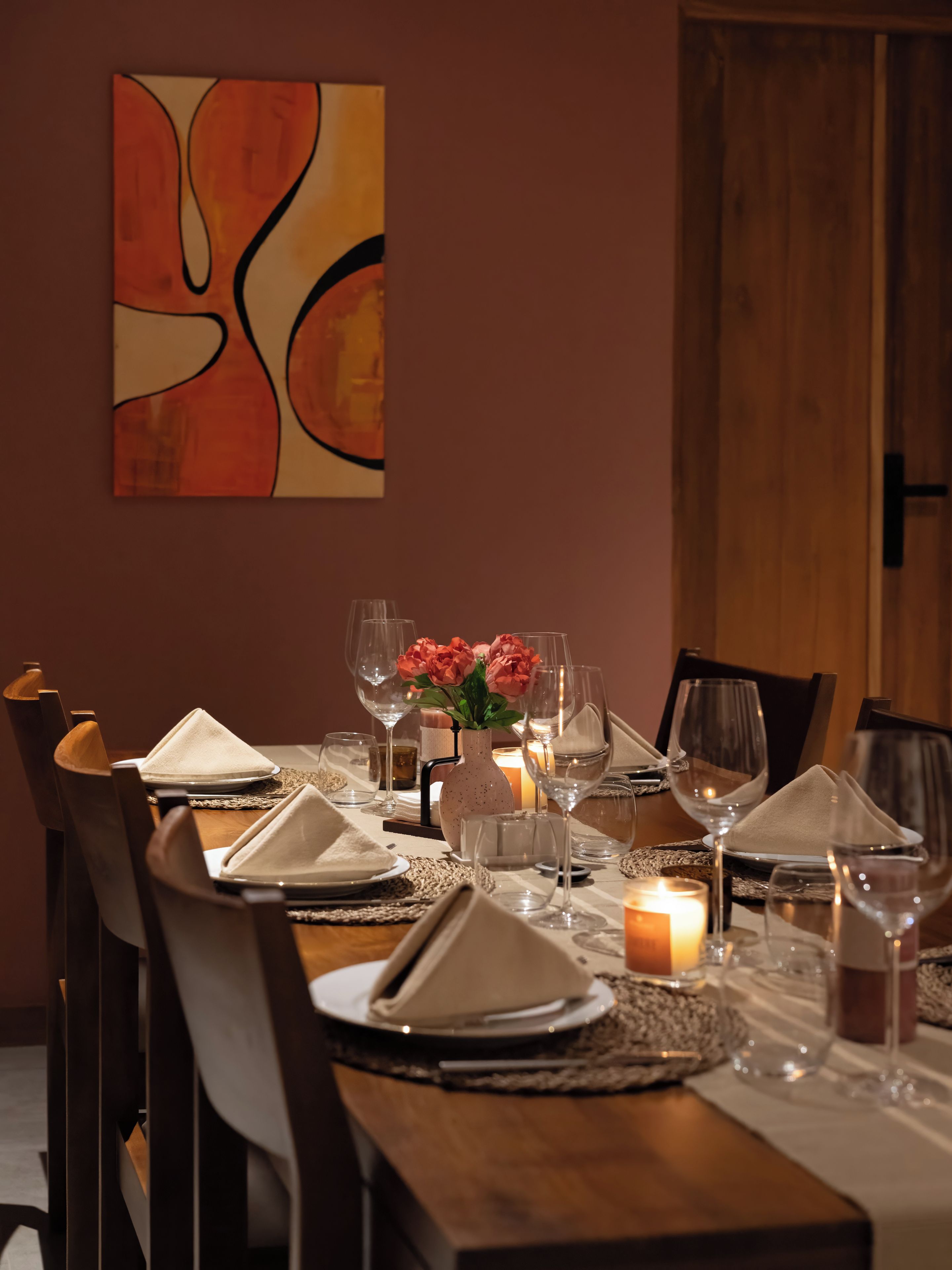 Una mesa de comedor dispuesta con platos, vasos y una vela, con un florero de flores y un cuadro en la pared.