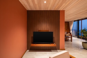 A modern living room with a large flat-screen TV mounted on a wooden wall, a dining area with a table and chairs, and a view of the outdoors through large windows.