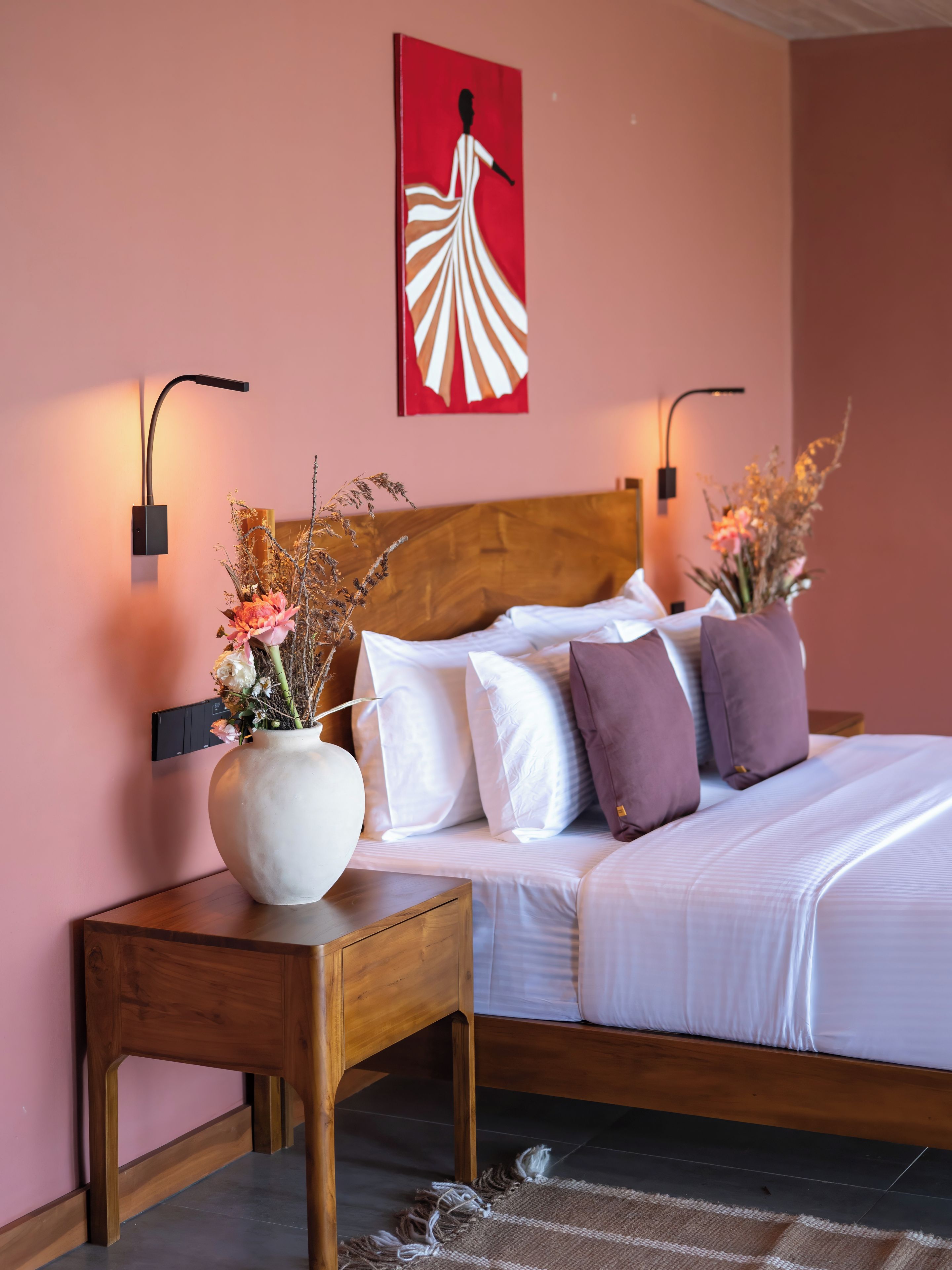 Quarto de hotel com cabeceira de madeira, roupa de cama branca, um vaso com flores secas e duas luminárias fixadas na parede.