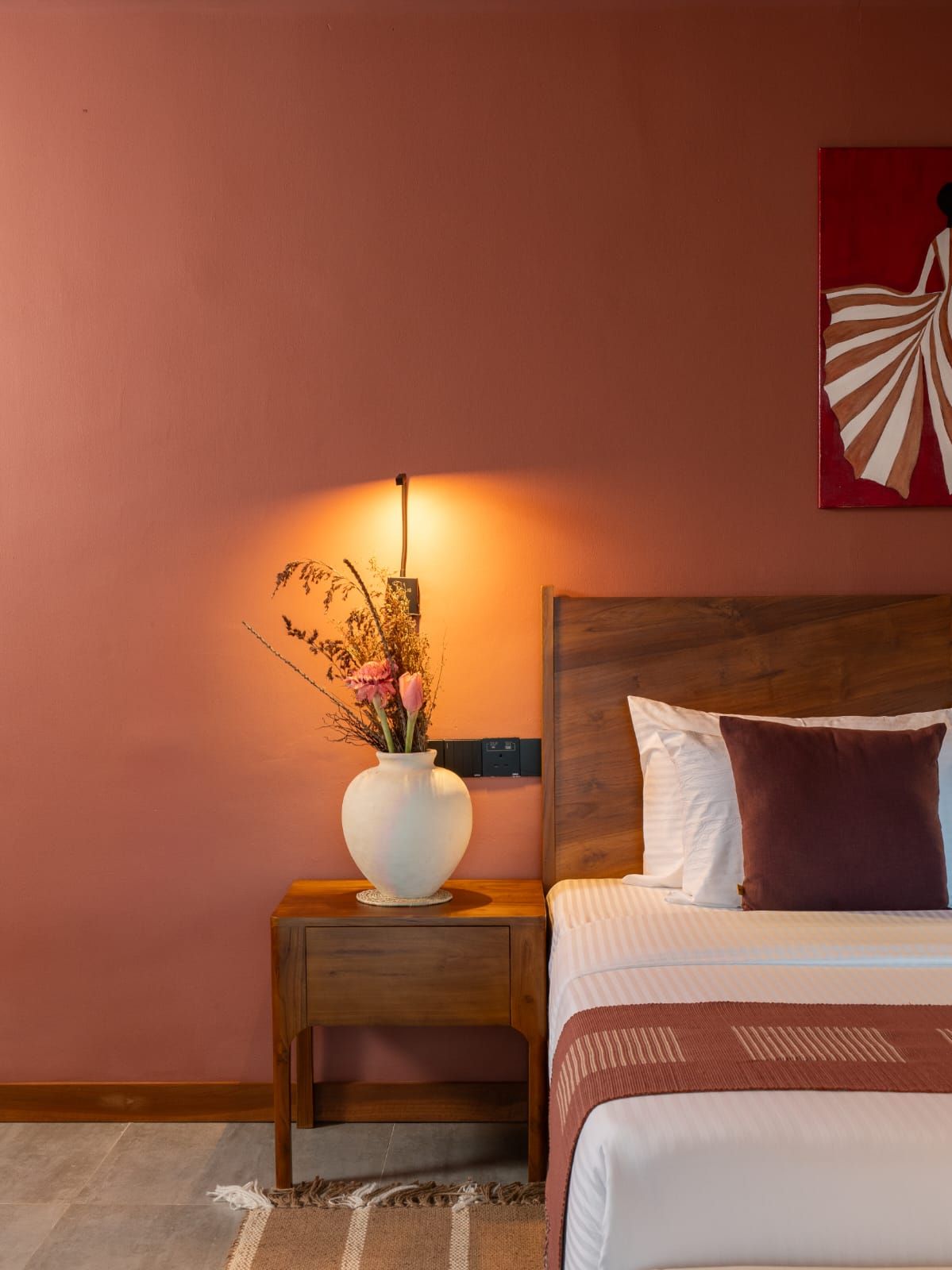 A bedroom with a wooden headboard, a bedside table with a vase of flowers, and a wall-mounted power outlet.