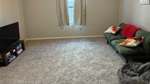 A living room with a flat-screen TV, a green sofa with floral cushions, a red throw blanket, and a window with beige curtains. -  New, clean, spacious and cozy! Pet friendly!  (Broken Arrow)