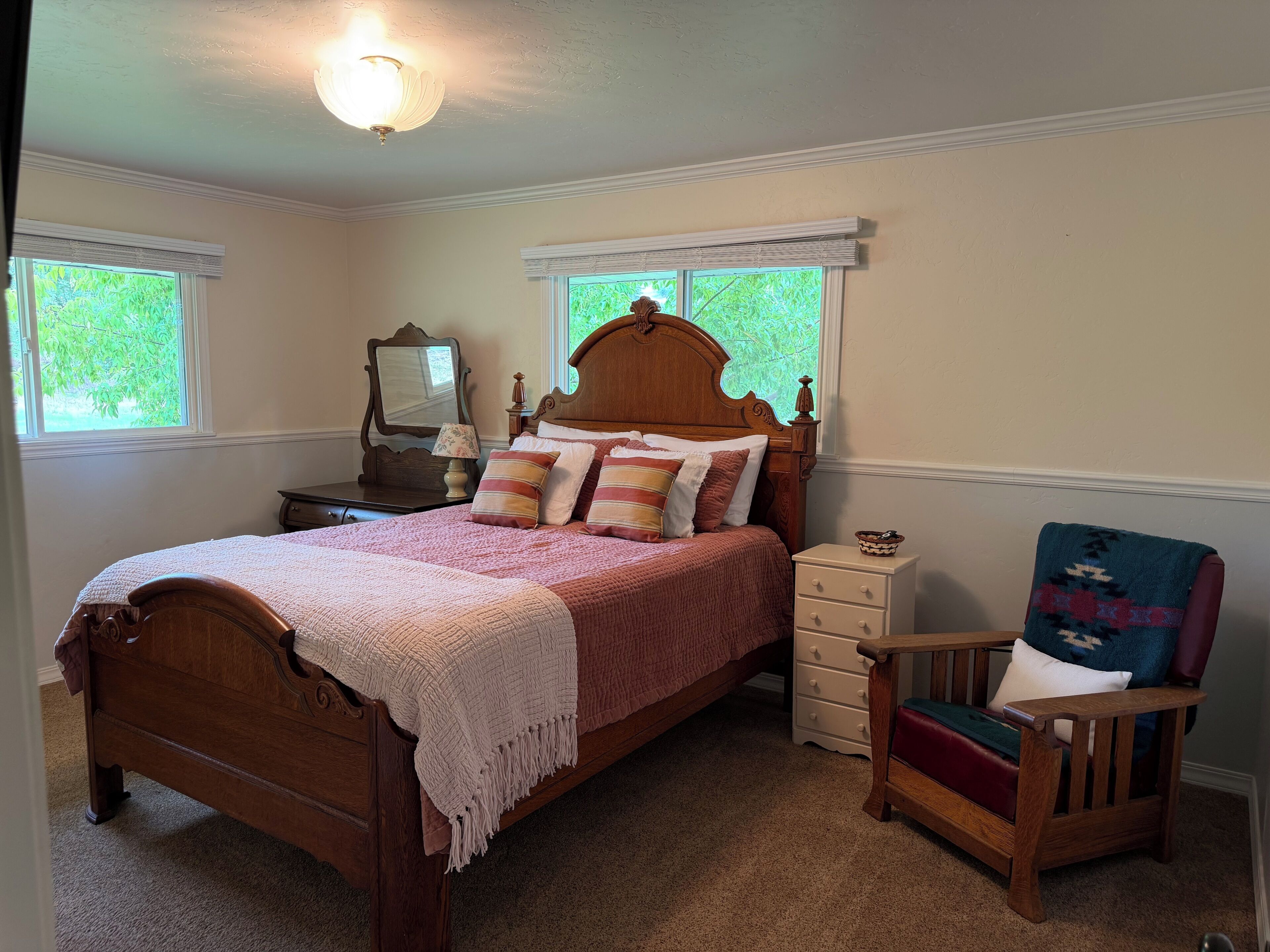 Une chambre à coucher avec un lit en bois, une table de chevet, une commode, une chaise et deux fenêtres équipées de stores.