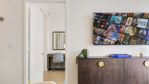 Ein Wohnzimmer mit an der Wand befestigtem Fernseher, der Filmplakate zeigt, einem Holzschrank mit runden Griffen und einem Sessel mit gemustertem Kissen.