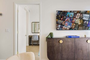 Ein Wohnzimmer mit an der Wand befestigtem Fernseher, der Filmplakate zeigt, einem Holzschrank mit runden Griffen und einem Sessel mit gemustertem Kissen.