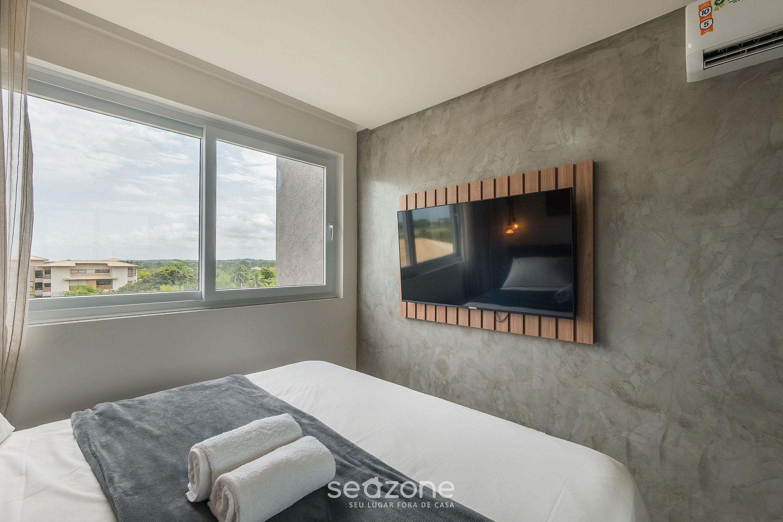 A hotel room with a large window, a flat-screen TV mounted on the wall, a bed with a grey blanket and two towels, and a textured grey wall.