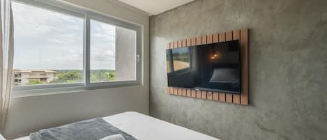 A hotel room with a large window, a flat-screen TV mounted on the wall, a bed with a grey blanket and two towels, and a textured grey wall.