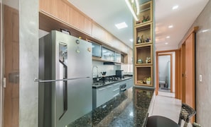 A modern kitchen with a built-in refrigerator, stove, and microwave. There is a wooden cabinet above the stove and a small shelf with decorative items. - Apart in Condo w/ Pool and BBQ Aga0304 (Camaçari)
