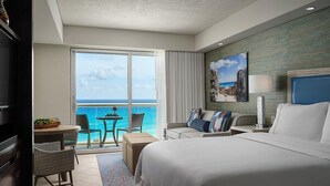 Habitación de hotel con una cama grande, un escritorio con una silla, una mesita con una silla y vista al océano a través de una puerta corrediza.