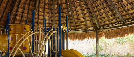 A playground with a thatched roof, a yellow slide, and climbing structures.