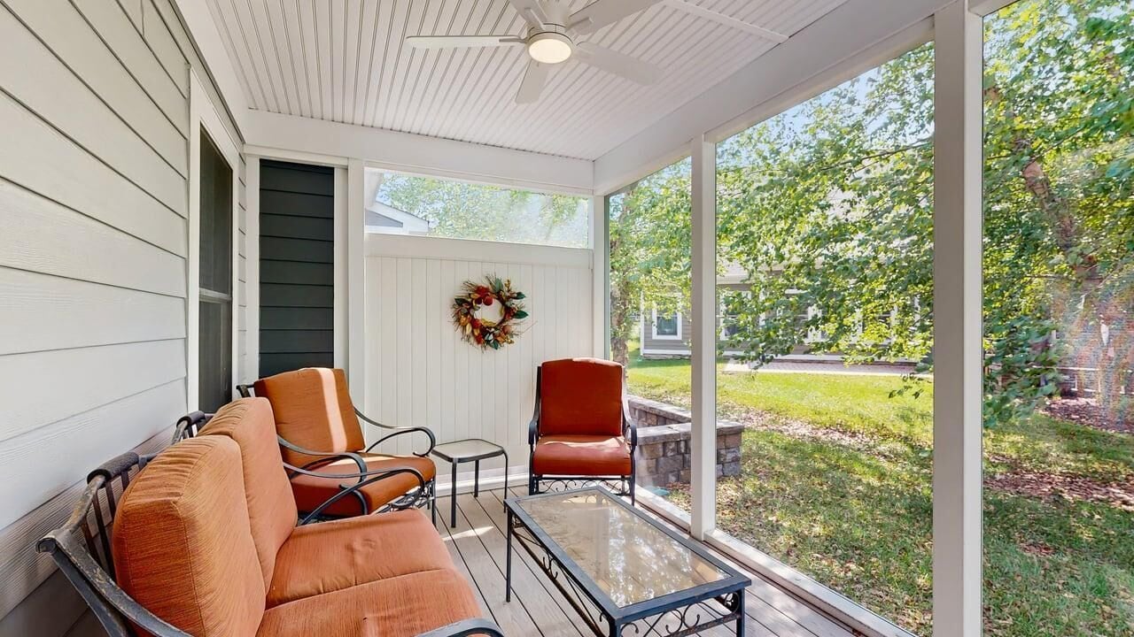 A covered porch with a glass table and orange chairs, a red chair, and a wreath on the wall.