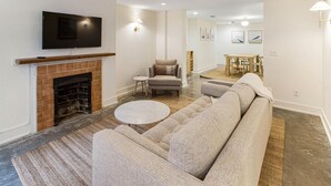 A modern living room with a fireplace, a flat-screen TV mounted above it, a sectional sofa, a round coffee table, and a dining area in the background. - Park House Garden - Ground Floor Two-Bedroom Apartment on Monterey Square (Savannah)