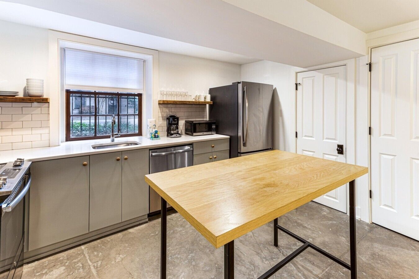 A modern kitchen with a wooden table, stainless steel appliances, and a window with blinds.