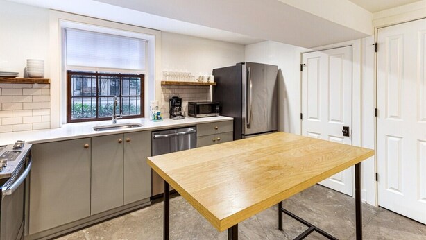 A modern kitchen with a wooden table, stainless steel appliances, and a window with blinds. - Park House Garden - Ground Floor Two-Bedroom Apartment on Monterey Square (Savannah)