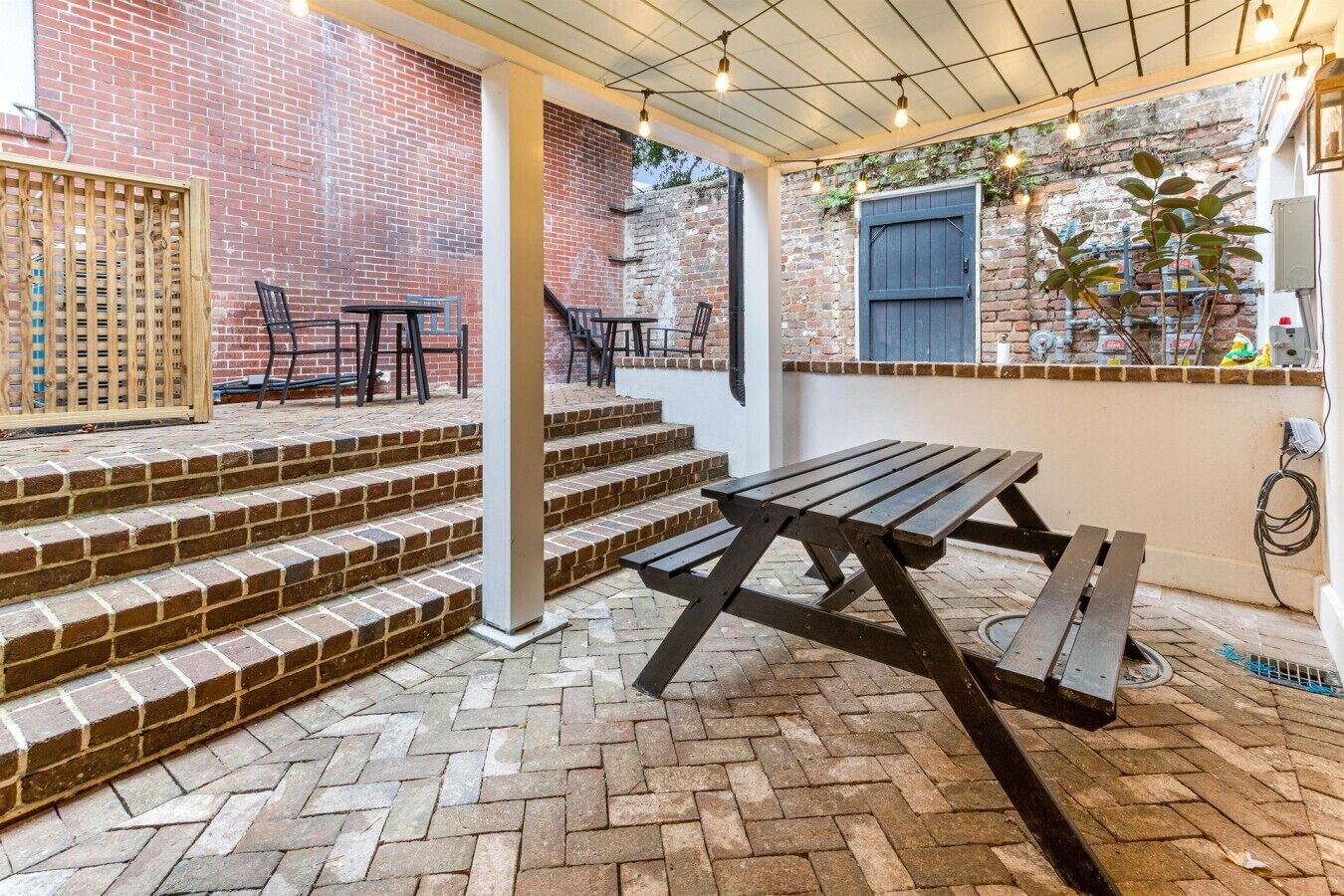 Une terrasse en briques avec une table de pique-nique et des chaises, des marches menant à un espace abrité doté d’un mur en briques et d’une fenêtre.