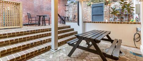Une terrasse en briques avec une table de pique-nique et des chaises, des marches menant à un espace abrité doté d’un mur en briques et d’une fenêtre.